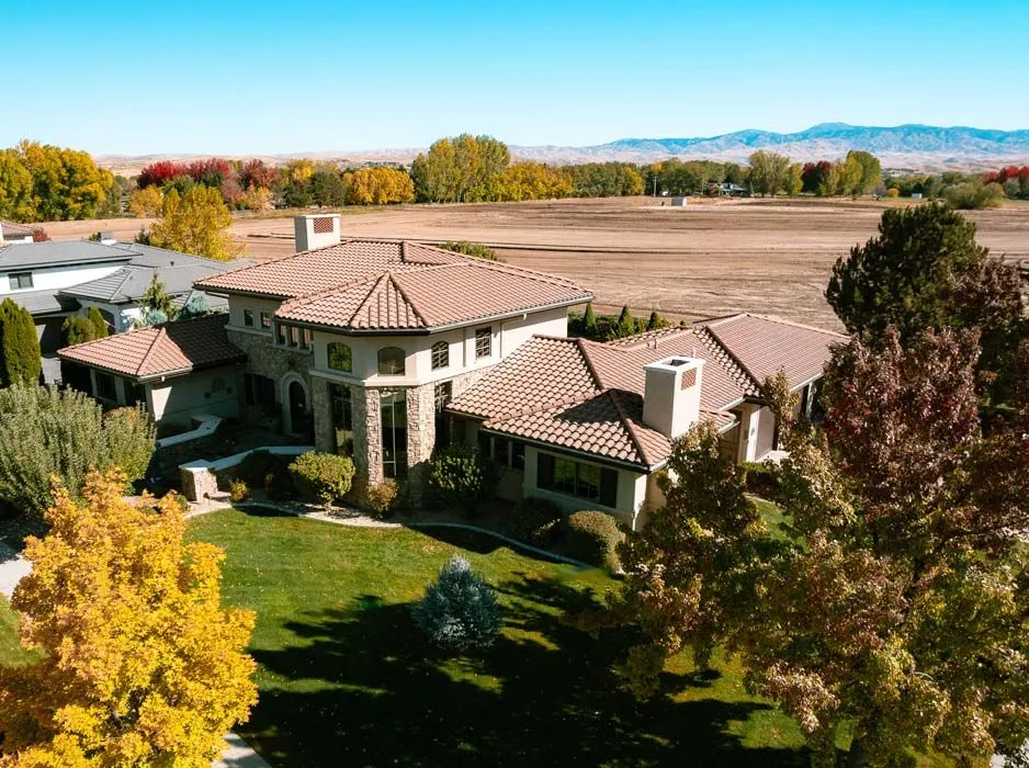 Aerial view of a large house with a tile roof and green lawn surrounded by colorful autumn trees and a field.