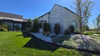 Modern house with gray brick facade, lush green lawn, and landscaping under a blue sky.