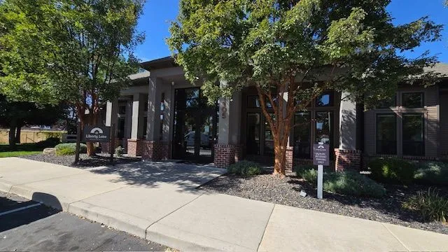 Building entrance with pillars, brown trim, and a sign under trees on a sunny day.