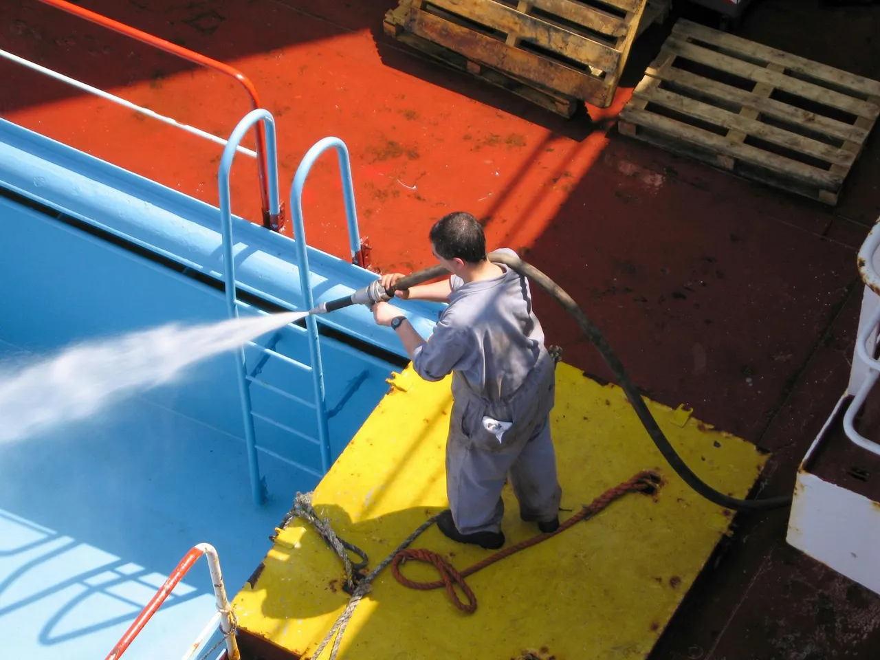 Person in blue coveralls sprays water into a blue pool on a red deck with wooden pallets nearby.