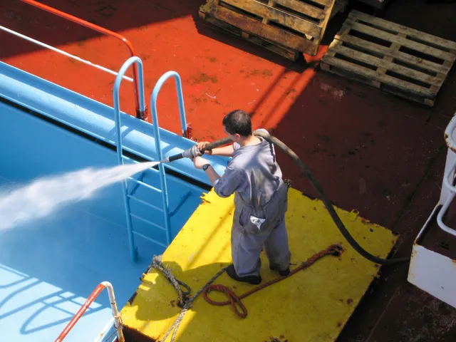 Person in blue coveralls sprays water into a blue pool on a red deck with wooden pallets nearby.