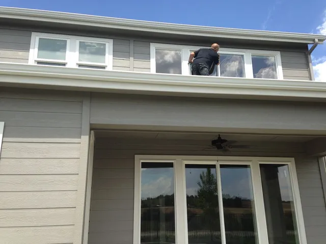 Person cleaning upper windows on a gray house. Sky and trees visible.