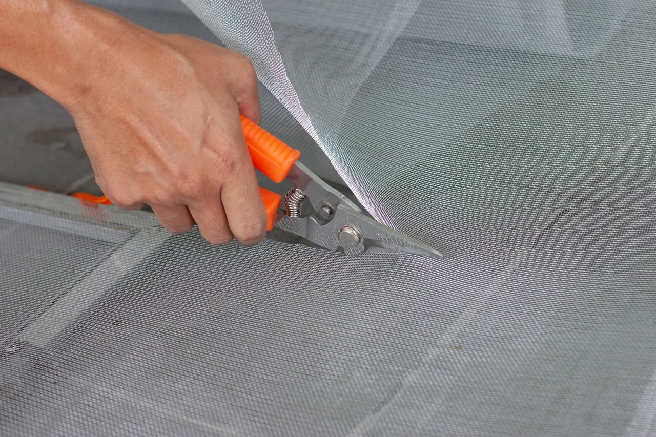 Hand using orange-handled scissors to cut silver mesh material.